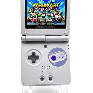 May include: A silver Game Boy Advance SP handheld gaming console with a clamshell design. The screen displays the Mario Kart Super Circuit title screen. The device features a black directional pad, two purple A and B buttons, and two small grey buttons.