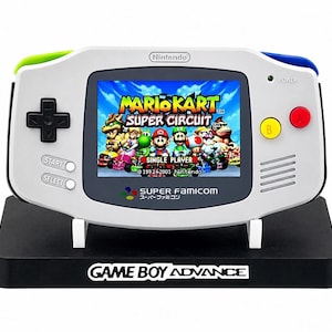 May include: A white handheld video game console with a green left shoulder button and blue right shoulder button. The screen displays the Mario Kart Super Circuit title screen. It features a black directional pad, yellow B button, red A button, and Start and Select buttons. The console rests on a black stand labelled Game Boy Advance.