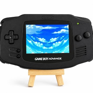 May include: A black Game Boy Advance handheld gaming console with a horizontal layout, featuring a directional pad, A and B buttons, Start and Select buttons, and a screen displaying a blue sky with white clouds. The Nintendo logo is visible.