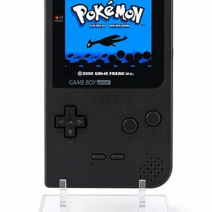 May include: A black Game Boy Pocket handheld gaming console featuring a screen displaying the Pokémon Silver Version title screen with the 2000 Game Freak Inc. copyright text. The device is mounted on a clear acrylic display stand with Game Boy text.