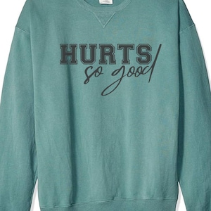 Op de afbeelding: Tealblauwe trui met de tekst "HURTS so good" in zwart.