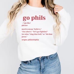 Może przedstawiać: Biała bluza z czerwonym tekstem, który brzmi "go phils /go filz/ phrase used to mean "believe," "rise above," "let's go fightins!" See also: "ring that bell," or "hit that jawn." origin: philadelphia"