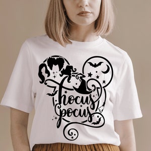 May include: White t-shirt with a black graphic design of the Sanderson sisters from the movie Hocus Pocus. The design includes a witch's hat, stars, a crescent moon, a bat, and a spider. The text "Hocus Pocus" is written in a stylized font.