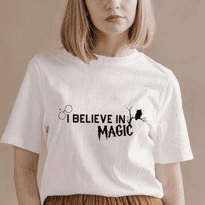 May include: White t-shirt with a black graphic that says "I believe in magic" with a lightning bolt and an owl on a branch. There are also two circles above the text.