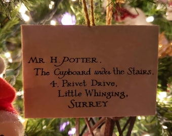 May include: A white card with black text hanging on a Christmas tree. The text reads: "MR. H. POTTER, The Cupboard under the Stairs, 4, Privet Drive, Little Whinging, SURREY".