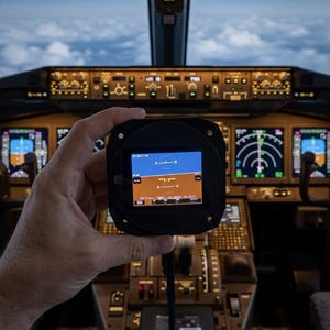 Op de afbeelding: Een handzaam digitaal vluchtinstrument met een blauw en bruin kunstmatige horizon-display en numerieke pitch-markeringen, gehouden voor de achtergrond van een vliegtuigcockpit met verlichte bedieningspanelen.