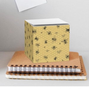 May include: A yellow cube-shaped notepad with a black and white pattern of bees and honeycombs. The notepad is sitting on top of a stack of notebooks.