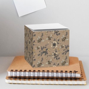 May include: A decorative cube notepad with a floral and bee pattern in shades of brown, gray, and cream. The notepad sits atop two spiral-bound notebooks. A small potted plant and a woven basket are visible in the background.
