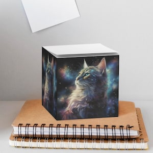 May include: A square note cube featuring a vibrant cosmic nebula background with a detailed illustration of a cat looking upward. The cat has soft fur and is surrounded by swirling colors of blue, purple, and gold, resembling a starry galaxy.