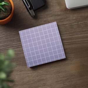 May include: A square, lavender sticky note with a white grid pattern sits on a wooden surface. A small potted plant, a pen, and a portion of a laptop are also visible. The note is a simple, functional office supply.