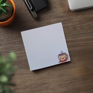 May include: A white sticky note with a cartoon coffee cup illustration in the corner. The coffee cup is smiling and has steam rising from it. A pen, stapler, small plant, and laptop are also visible on a wooden surface.