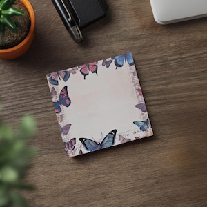 May include: Square notepad with a butterfly design. The notepad has a border of colourful butterflies in shades of blue, pink, and purple. The background is a soft cream colour with faint text. A pen and small plant are in the background.