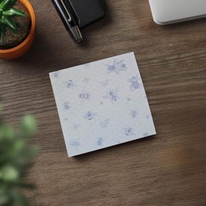 May include: Square sticky note pad with a light blue bee and honeycomb pattern. The pad is on a wooden surface, with a small potted plant, a pen, and a laptop visible in the background.