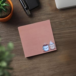 May include: A square, dusty rose sticky note pad with a cartoon illustration of two smiling coffee cups. One cup is blue with chocolate, the other is beige with whipped cream. The pad is on a wooden surface.