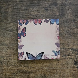 May include: A white notepad with a border of colorful butterflies. The butterflies are in shades of blue, red, and brown. The notepad has a blank white space in the center.