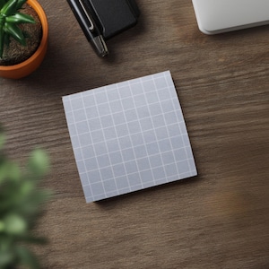 May include: A square, light gray sticky note with a white grid pattern. The note is on a wooden surface, with a small potted plant, a pen, and a portion of a laptop visible in the background. The overall theme is office supplies.