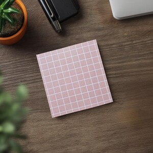 May include: A square, pink sticky note with a white grid pattern rests on a wooden surface. A small potted plant, a pen, and a portion of a laptop are also visible. The scene suggests a workspace or desk setting.