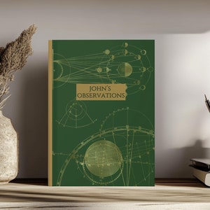 May include: A dark green hardcover journal featuring gold-colored celestial diagrams and the text "JOHN'S OBSERVATIONS" printed in a rectangular box on the front cover. The book has a gold-colored spine and a clean, vertical orientation.