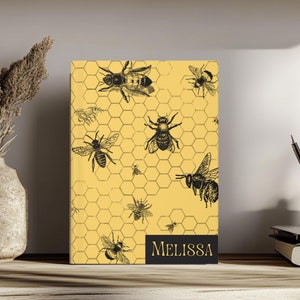 May include: A yellow notebook with a honeycomb pattern and black bee illustrations. The notebook has the name "Melissa" printed on the bottom.