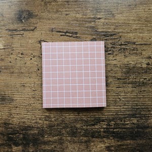 May include: A square, pink sticky note pad with a white grid pattern. The notepad is centered on a dark brown, textured wooden surface.
