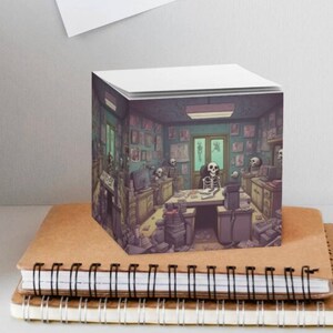 May include: A cube-shaped notepad with a printed image of a room with a skeleton sitting at a table. The room is filled with various objects, including a computer, a lamp, and a clock. The notepad is sitting on top of a stack of notebooks.