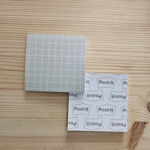 May include: Two square Post-it note pads on a wooden surface. One pad is light grey with a white grid pattern. The other pad is white with the Post-it logo repeated across the surface. The background is a light brown wood.
