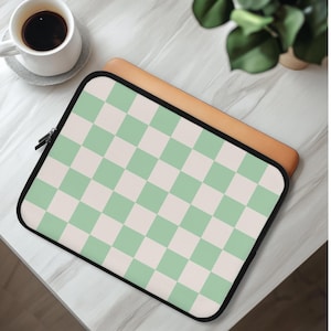 May include: A laptop sleeve with a mint green and white checkered pattern. The sleeve has a black zipper and a black border. A cup of coffee and a plant are in the background.