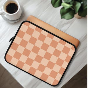 Puede incluir: Una funda para portátil con un patrón de tablero de ajedrez melocotón y crema. La funda tiene una cremallera y un ribete negros. Una taza de café está sobre un platillo blanco en la esquina superior izquierda de la imagen.