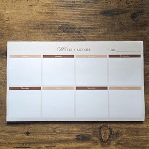 May include: A white weekly agenda notepad against a wooden background. The notepad features the words "WEEKLY AGENDA" in script. Each day of the week is labelled with a brown and tan border, with space for notes.