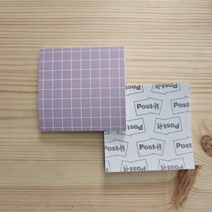 May include: Two square Post-it notes on a wooden surface. One is lavender with a white grid pattern. The other is white with the Post-it logo in grey. Office supplies.