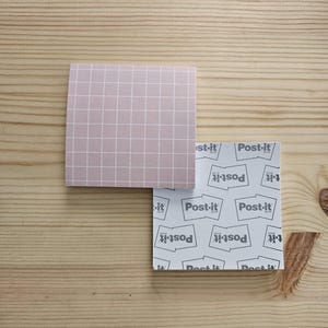 May include: Two Post-it note pads on a wooden surface. One pad is pale pink with a white grid pattern. The other pad is white with a repeating Post-it logo. The Post-it pads are square-shaped.