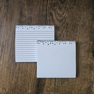 May include: Two white notepads with black decorative borders and a wooden background. One notepad has horizontal lines, and the other is blank. The borders feature a repeating pattern of small stars and lines.