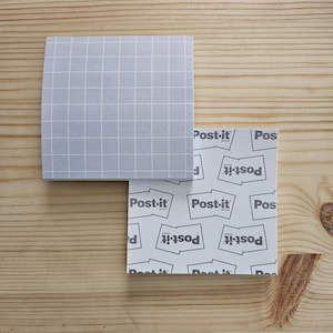 May include: Two square Post-it note pads. One pad is light gray with a white grid pattern. The other pad has a white background with the Post-it brand name printed repeatedly in gray. The pads are on a light brown wooden surface.