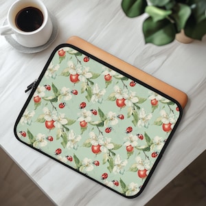 Może przedstawiać: Prostokątne etui na laptopa z jasnym szałwiowo-zielonym tłem i powtarzającym się wzorem białych kwiatów jabłoni, zielonych liści, czerwonych jabłek oraz małych czerwonych biedronek w czarne kropki. Etui posiada czarny zamek błyskawiczny i wykończenie.