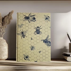 Op de afbeelding: Een geel notitieboek met een honingraatpatroon en blauwe bijenillustraties. Het notitieboek ligt op een houten oppervlak met een vaas met gedroogde bloemen en een pennenhouder met pennen.