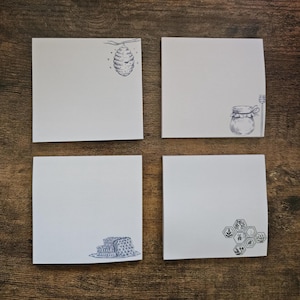May include: Four white square notepads with blue ink illustrations of a beehive, honey jar, honeycomb, and a stack of honeycombs. The notepads are arranged on a wooden surface.