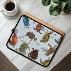 May include: A light blue laptop sleeve with a pattern of colourful sea turtles, coral, and seashells. The sleeve has a black zip and a tan leather-like border. A white coffee cup sits on a grey coaster.