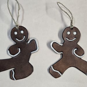 May include: Two brown gingerbread man decorations with white icing details and string hangers. Each decoration has a smiling face and white accents on the arms and legs. The decorations are designed to look like they are dancing.