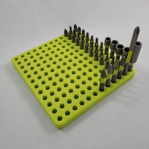 May include: A lime green, hexagonal-holed organizer holding various sizes of screwdriver bits. The bits are silver and black, with some having yellow accents. The organizer is square and made of plastic.