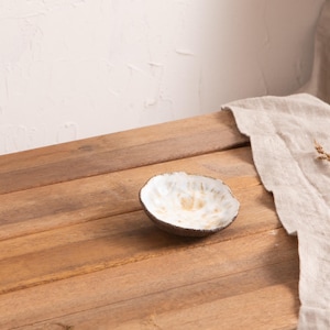 Könnte beinhalten: Eine kleine, handgefertigte Keramikschale mit strukturiertem, cremefarbenem Innenraum und dunklem braunen Rand. Die Schale steht auf einer Holzoberfläche, mit einem neutralfarbenen Leinentuch und getrockneten floralen Elementen im Hintergrund.