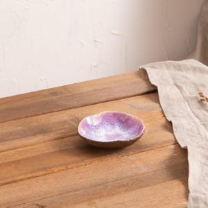 Könnte beinhalten: Eine kleine, handgefertigte Keramikschale mit einer einzigartigen, organischen Form. Das Innere zeigt einen Farbverlauf von Lila und Weiß, während die Außenseite eine natürliche, erdige braune Farbe hat. Die Schale steht auf einer Holzoberfläche.
