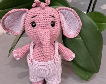 Può includere: Un giocattolo di elefante rosa all'uncinetto che indossa una salopette lilla chiaro con due bottoni bianchi. L'elefante ha un piccolo naso rosa e occhi neri.