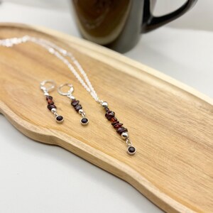May include: A silver necklace and matching earrings set. The necklace features a strand of small, dark red gemstones and silver beads. The earrings have a similar design, with dark red gemstones and silver accents. The jewelry is displayed on a wooden tray.