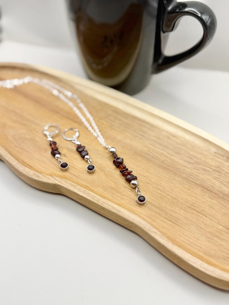 May include: A silver necklace and matching earrings set. The necklace features a strand of small, dark red gemstones and silver beads. The earrings have a similar design, with dark red gemstones and silver accents. The jewelry is displayed on a wooden tray.