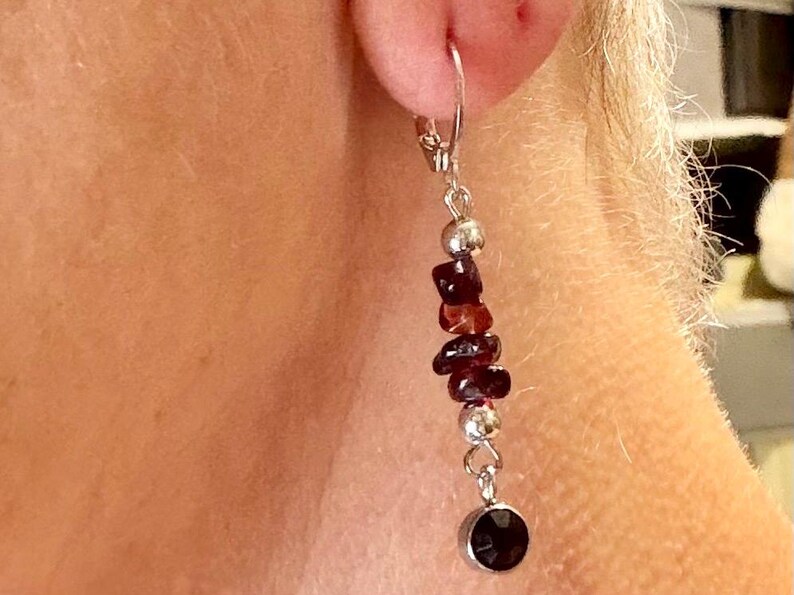 May include: Dangling earring with a silver-tone metal hook and a cascade of dark red-brown stones. The stones are interspersed with silver beads, and the earring ends with a dark red-brown, round stone. The earring is shown against a person's skin.