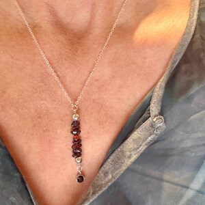 May include: A silver necklace with a vertical pendant featuring a series of dark red garnet stones and small silver beads. The necklace is worn against a person's skin, with a blue and gray top in the background.