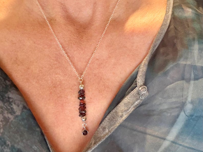 May include: A silver necklace with a vertical pendant featuring a series of dark red garnet stones and small silver beads. The necklace is worn against a person's skin, with a blue and gray top in the background.