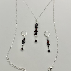May include: A silver necklace and earring set featuring dark red gemstone beads. The necklace has a pendant with a vertical arrangement of beads, and the earrings have a similar design. The set includes a silver chain and earring hooks.