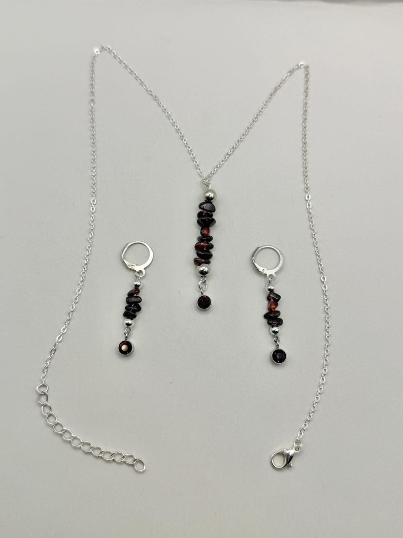 May include: A silver necklace and earring set featuring dark red gemstone beads. The necklace has a pendant with a vertical arrangement of beads, and the earrings have a similar design. The set includes a silver chain and earring hooks.