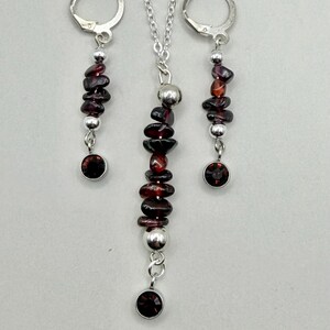 May include: A jewelry set featuring a necklace and a pair of earrings. The necklace has a silver chain with a pendant of stacked, dark red gemstones and silver beads, and a single red gemstone at the bottom. The earrings have a similar design.